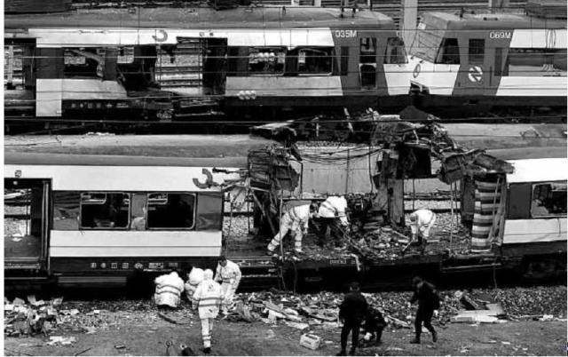 Agentes policiales buscan entre los restos, mientras los cochas 3 y 4 del tren de Atocha pasan por detrás. Son aproximadamente las 13 h